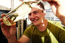 Ein junger Mann hält eine Flasche mit hellem Inhalt gegen das Licht. © Hessen schafft Wissen / Daniel Chassein