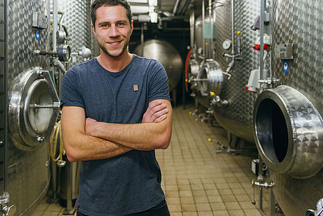 Ein junger Mann im blauen T-Shirt steht vor Edelstahlfässern in einem Weinkeller © Hessen schafft Wissen – Steffen Böttcher