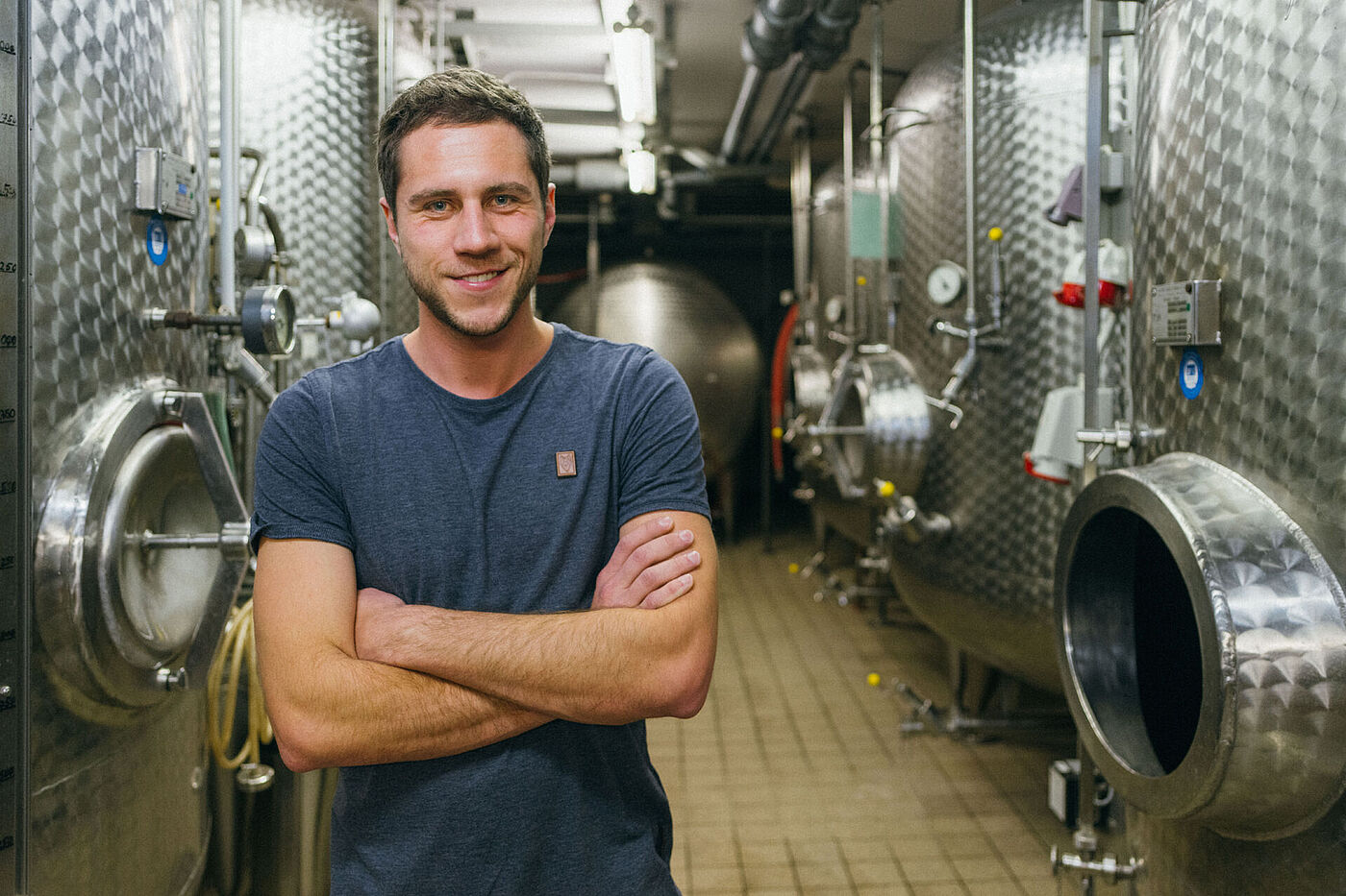 Ein junger Mann im blauen T-Shirt steht vor Edelstahlfässern in einem Weinkeller © Hessen schafft Wissen – Steffen Böttcher