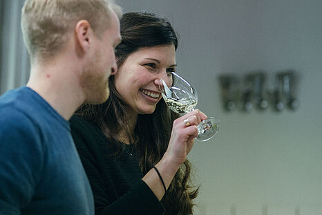 Eine junge Frau mit dunklen Haaren riecht an einem Glas mit Wein, ein blonder junger Mann steht daneben © Hessen schafft Wissen – Steffen Böttcher